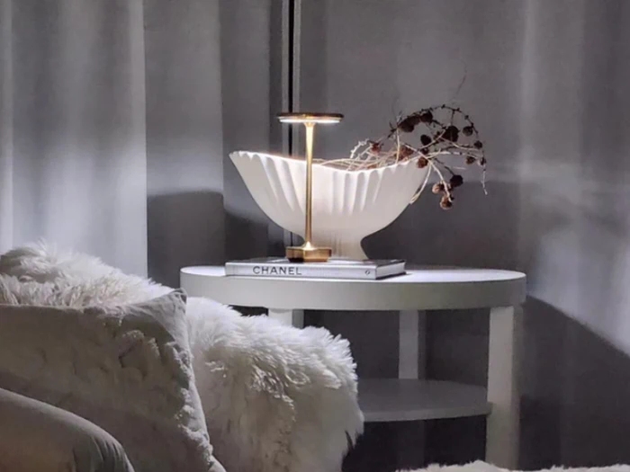 Et rundt hvidt sidebord med en hvid tekstureret lampe, en dekorativ gren og en Chanel-bog står ved siden af en blød hvid stol i et svagt oplyst rum med grå gardiner.