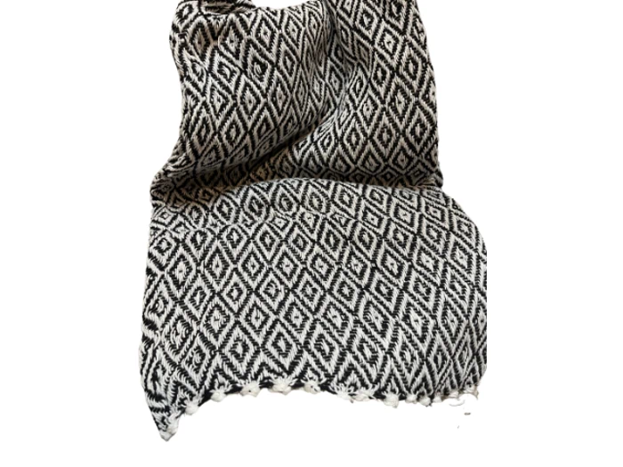 Et sort og hvidt vævet tekstil med et gentaget diamantmønster og frynsede kanter, lagt på en hvid baggrund.