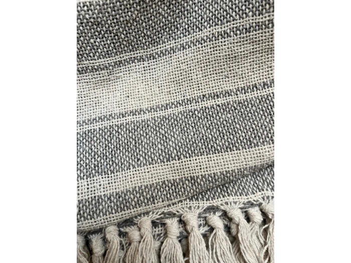 Nærbillede af et vævet tekstil med grå og hvide tråde, vandrette striber og frynser langs den nederste kant.