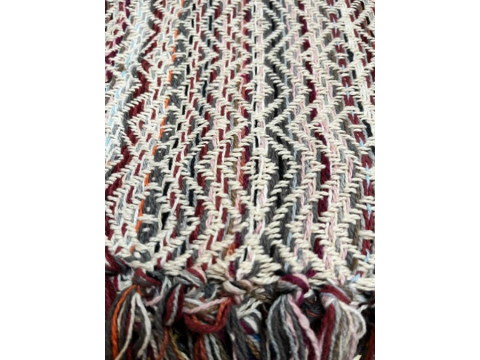 Nærbillede af et vævet tekstil med zigzagmønster i cremefarvede, grå, rødbrune og sorte farver, der ender i flerfarvede frynser i bunden.