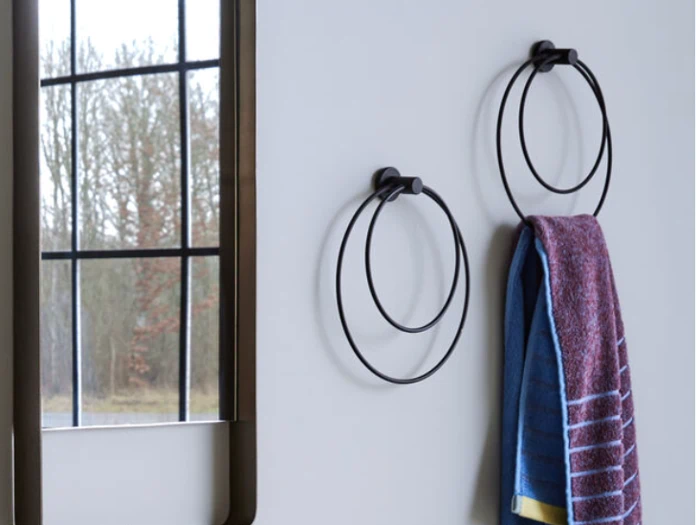 En badeværelsesvæg med to sorte håndklæderinge, hvoraf den ene holder et blåt og lilla håndklæde. Et vindue med udsigt til træer reflekteres i et spejl ved siden af håndklæderingene.