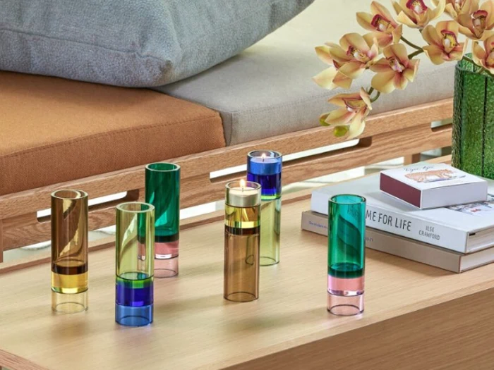 Seks farverige glasvaser står på et træbord ved siden af bøger og en grøn vase med gule orkideer; en sofa står i baggrunden.