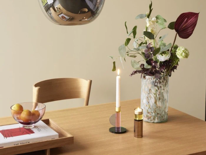 Et træbord med en vase med blomster, et tændt stearinlys, en skål med frugt og en bakke med en bog og et glas, og en træstol ved siden af.