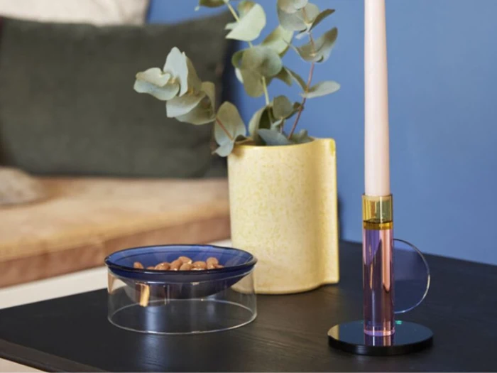 På et lille bord står en skål med nødder, en gul vase med eukalyptusgrene og et lyserødt stearinlys i en glasholder med en sofa og en blå væg i baggrunden.