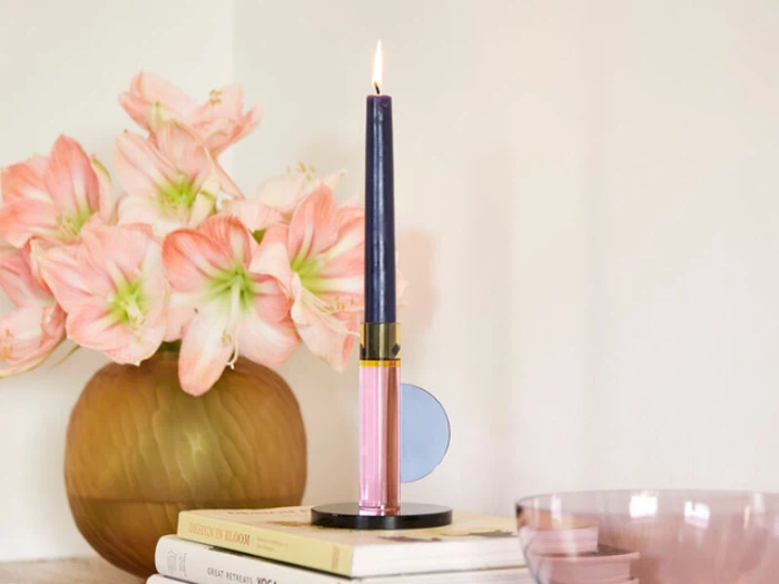 Et tændt marineblåt fyrfadslys står i en moderne holder på en stak bøger med en vase med lyserøde blomster i baggrunden.