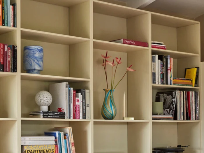 En beige bogreol fyldt med bøger, en blå marmorvase, en kugleformet lampe, en høj blå vase med lyserøde blomster, et stearinlys og forskellige pyntegenstande.