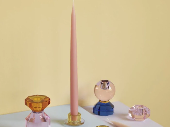 Et rosa fyrfadslys i en facetteret holder, omgivet af farverige, ædelstenslignende glaslysestager på en hvid overflade mod en lysegul baggrund.