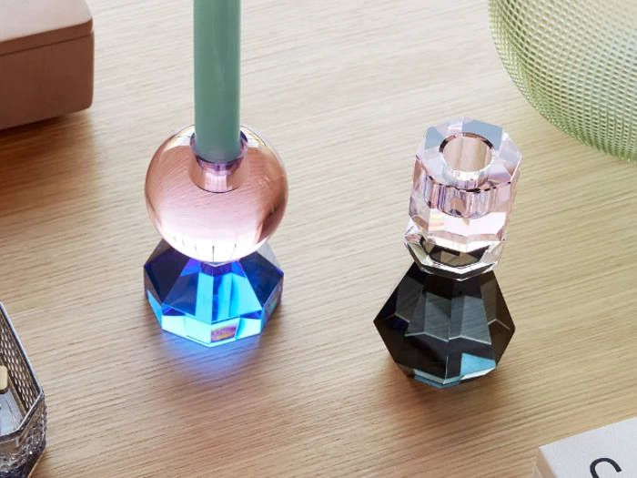 To facetterede glaslysestager, en blå og pink med et grønt lys og en klar og sort, står på en lys træplade blandt andre husholdningsartikler.