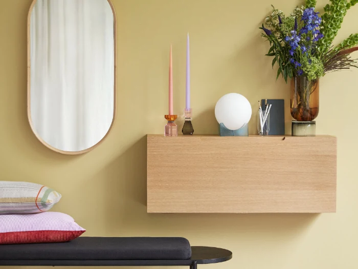 Et moderne interiør med et rundt spejl, en svævehylde i træ med stearinlys, en globelampe og en vase med blomster ved siden af en polstret bænk med farverige puder.