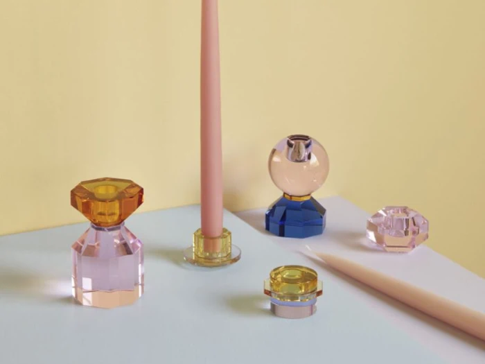 Fem farverige, geometriske glaslysestager holder pastelfarvede stearinlys på en lyseblå og gul baggrund i en blød belysning.