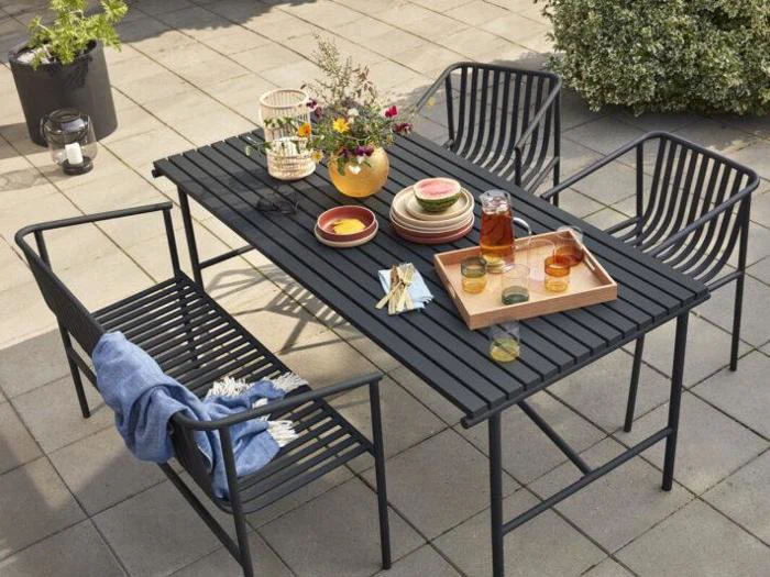 Et sort udendørs metalbord med matchende stole, dækket med tallerkener, glas, en gul vase med blomster og en bakke med drikkevarer på en terrasse. Et blåt tæppe er trukket over den ene stol.