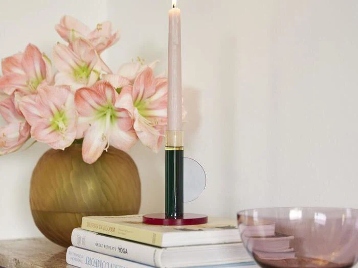 Et tændt lyserødt stearinlys står i en moderne holder på en stak bøger ved siden af en vase med lyserøde blomster og en gennemsigtig lyserød skål.