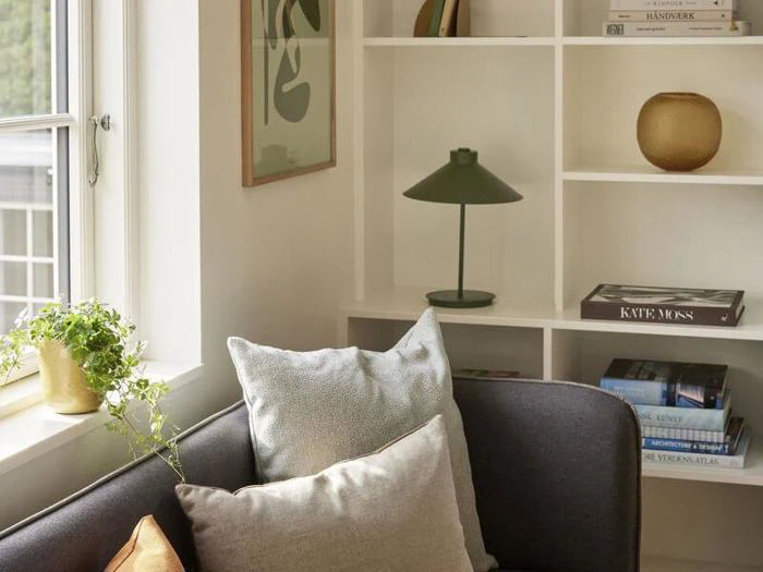 Et hyggeligt stuehjørne med en grå sofa, lyse puder, en grøn potteplante ved vinduet og hylder med bøger, en lampe og pyntegenstande.
