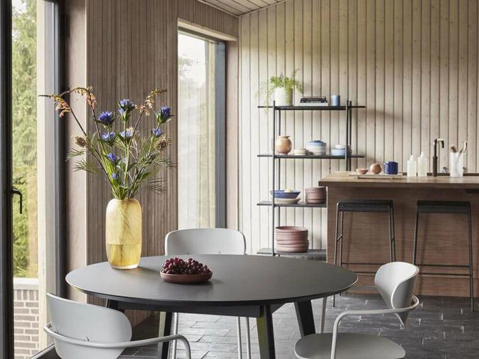 Et moderne køkken og spiseplads med et rundt sort bord, hvide stole, en gul vase med blomster og åbne hylder med service i baggrunden.