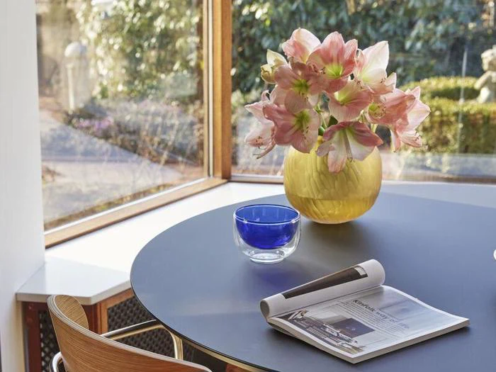 På et rundt bord ved et vindue står en gul vase med lyserøde blomster, en blå glasskål og en sammenrullet avis. Sollyset strømmer ind gennem haven udenfor.