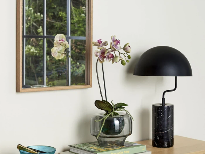 En glasvase med orkideer, en sort kuppellampe og bøger står på et træbord ved siden af et vægspejl, der reflekterer grønne planter.