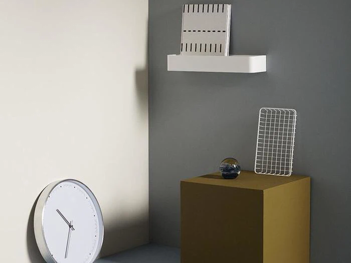 Et minimalistisk rum med et vægur, en hvid hylde med en slidset genstand, en gul piedestal med en glaskugle og et metalgitter, alt sammen mod grå og beige vægge.