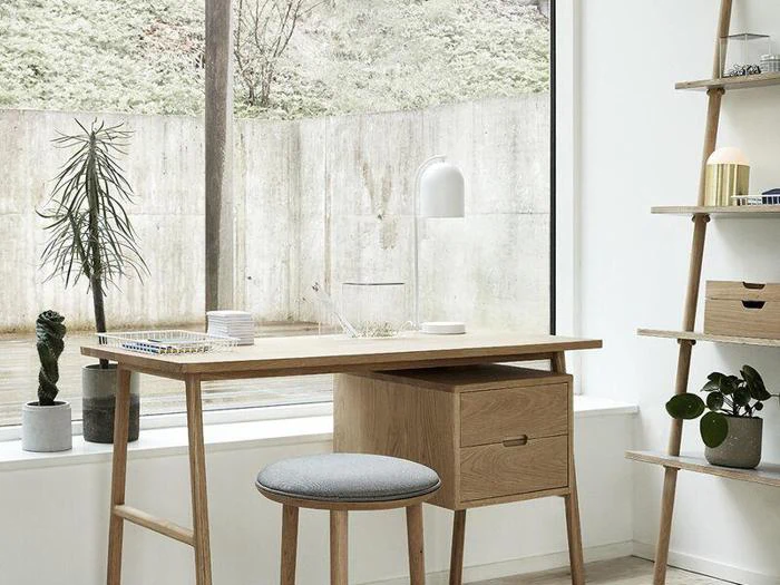 Et minimalistisk træskrivebord med en grå skammel, en hvid lampe og indendørs planter ved et stort vindue med udsigt til en betonvæg og grønne områder.