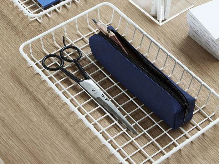 Et blåt penalhus og en sort saks placeret i en hvid trådkurv på en træoverflade.