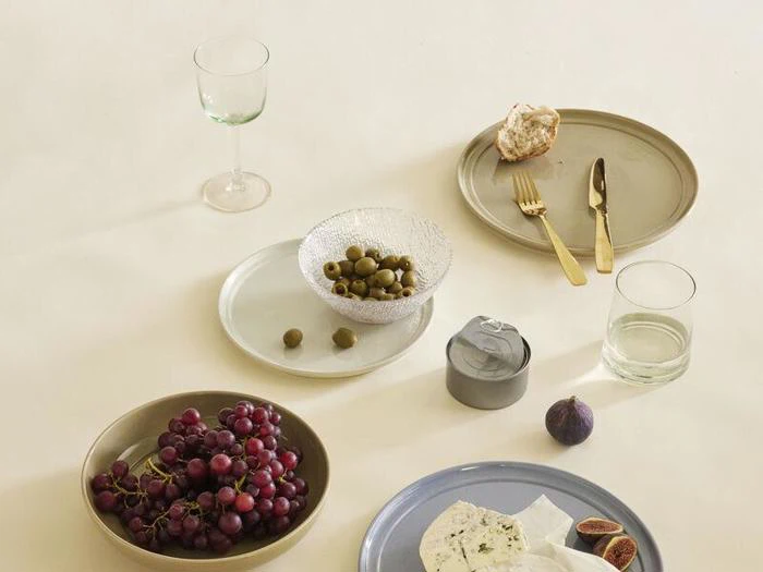Minimalistisk borddækning med vindruer, oliven, figner, ost, brød, et glas vin, vand, en dåse og guldbestik på neutrale tallerkener mod en lys baggrund.