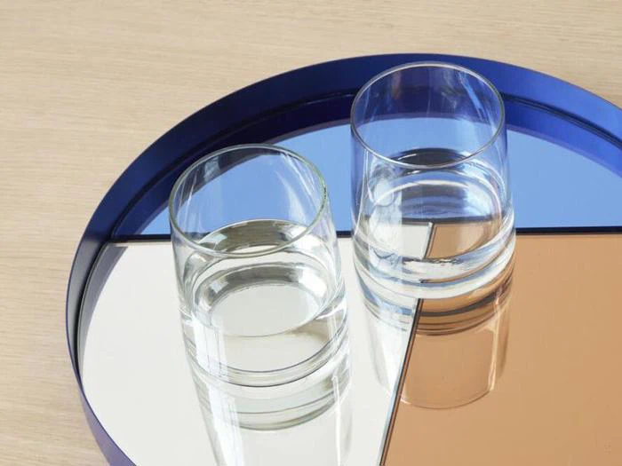 To klare vandglas står på en rund, flerfarvet bakke med blå, hvide og bronzefarvede sektioner, der er placeret på en lys træoverflade.