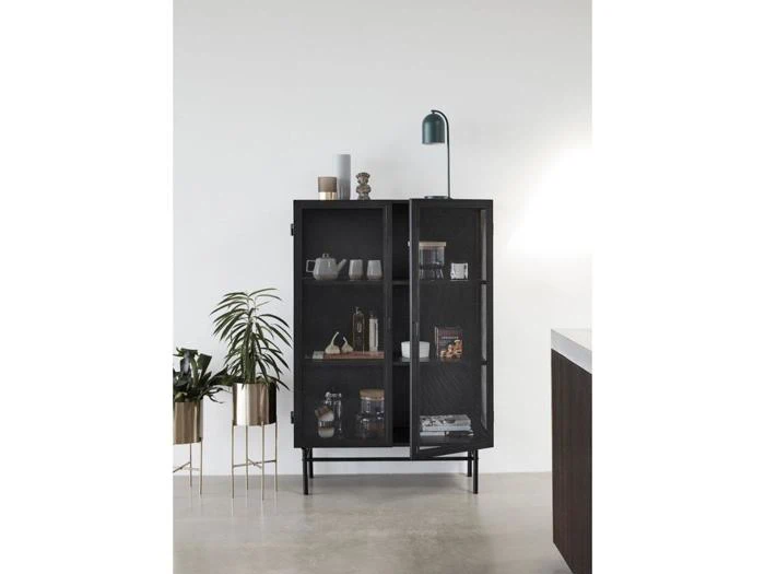 Et sort metalskab med glasdøre rummer indretnings- og køkkenting; en grøn plante i guldkrukker og en grøn skrivebordslampe står ved siden af mod en hvid væg og et betongulv.