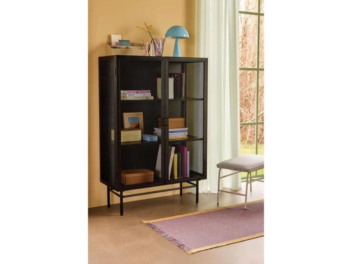 Et sort metal- og glasskab med hylder til bøger og indretning står ved et vindue, ved siden af en bænk og et lilla tæppe, i et blødt oplyst rum med beige vægge.