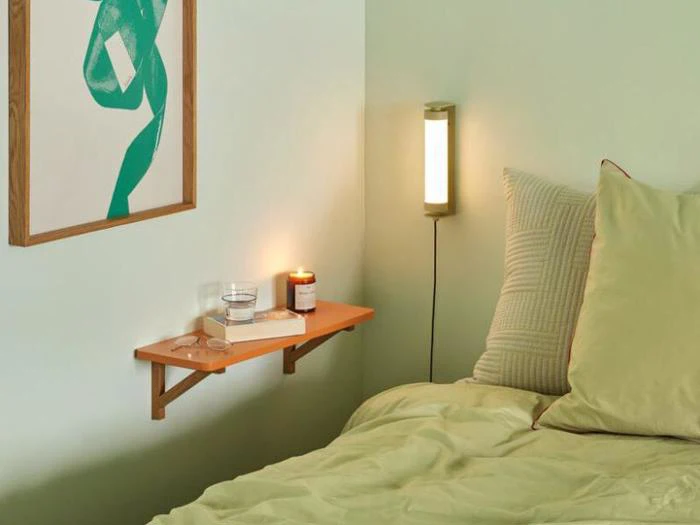 En pænt redt seng med grønt sengetøj ved siden af en væghylde med et stearinlys, et glas vand, briller og en bog, med en væglampe og abstrakt kunst over.