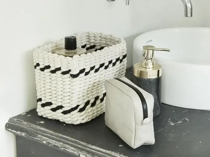 En flettet kurv, en lille toilettaske og en sæbedispenser er placeret på en mørk bordplade ved siden af en hvid vask.