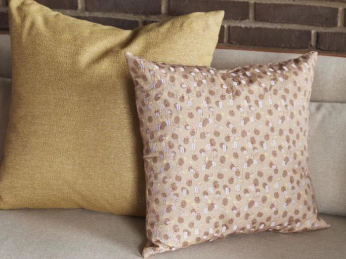 To firkantede puder på en beige sofa; den ene pude er ensfarvet gul, den anden er lysebrun med et prikmønster. I baggrunden ses en murstensvæg.