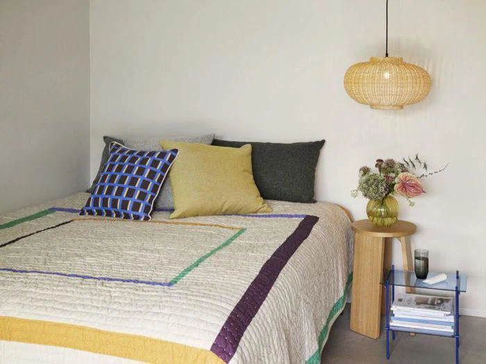 En pænt redt seng med farverige puder, en dyne med geometriske mønstre, et sidebord i træ med en lampe, en vase med blomster og en blå hylde med bøger under.