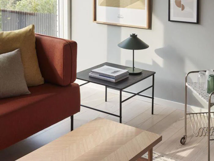 En moderne stue med en rustfarvet sofa, neutrale puder, et sort sofabord med bøger og en lampe, kunstværker på væggen og en lille rullevogn ved siden af.