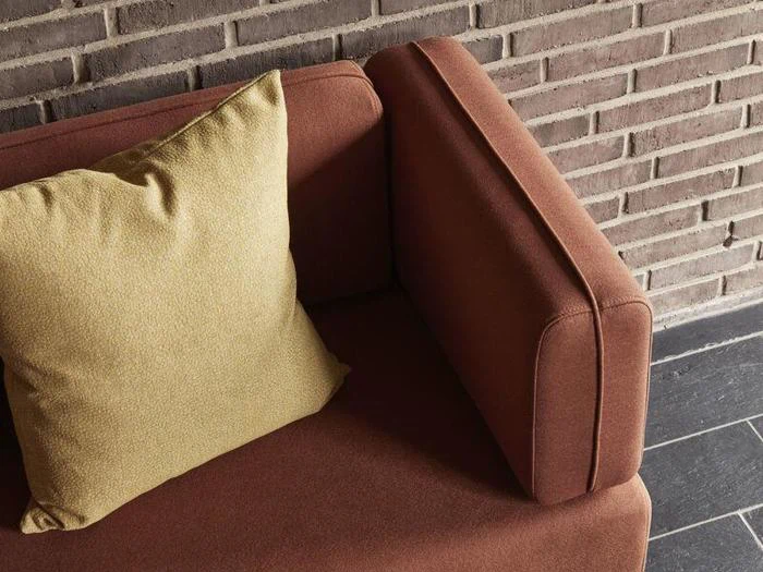 En brun sofa med en lysegul pude er placeret mod en murstensvæg og et mørkt flisegulv.