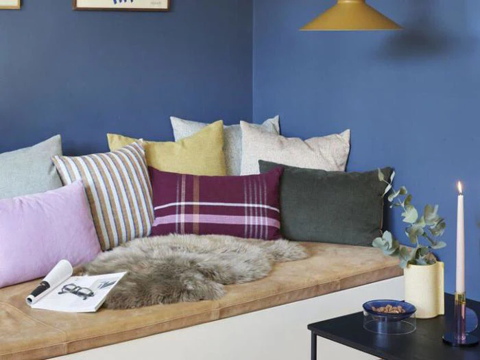 En hyggelig bænk med en solbrun pude, forskellige farverige puder, en pelskvast og et bord med et tændt stearinlys, en plante og en skål mod en blå væg.