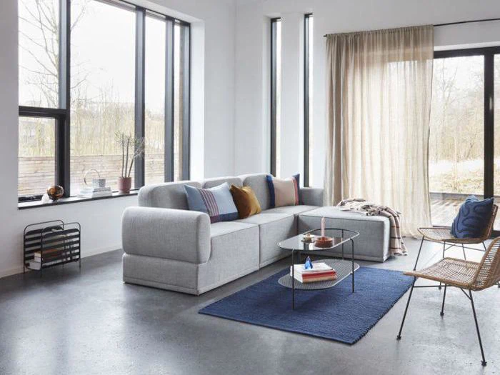 Moderne stue med en grå sektionssofa, blåt tæppe, sofabord i glas, kurvestole og store vinduer med gennemsigtige gardiner, der lukker naturligt lys ind.