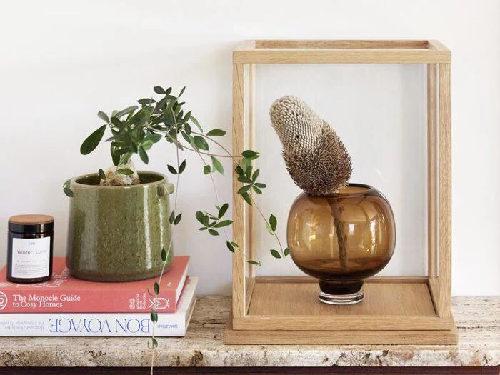 En grøn potteplante og en brun vase med en tørret blomst i en klar montre står på bøger oven på en rustik overflade med et stearinlys i nærheden.