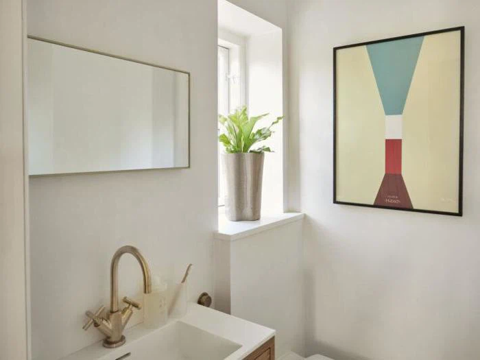 Et moderne badeværelse med et rektangulært spejl, potteplante i vindueskarmen, abstrakt vægkunst og en vask med guldarmatur.