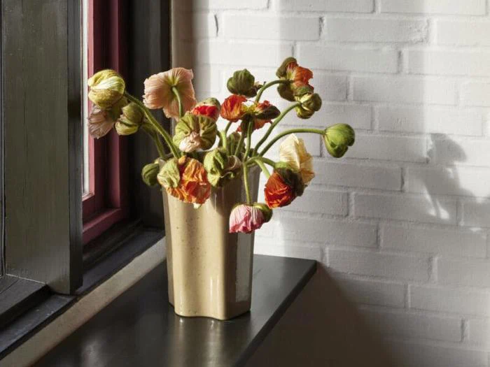 En beige vase med visne orange og gule valmuer står i en mørk vindueskarm mod en hvid murstensvæg, hvor sollyset kaster skygger.