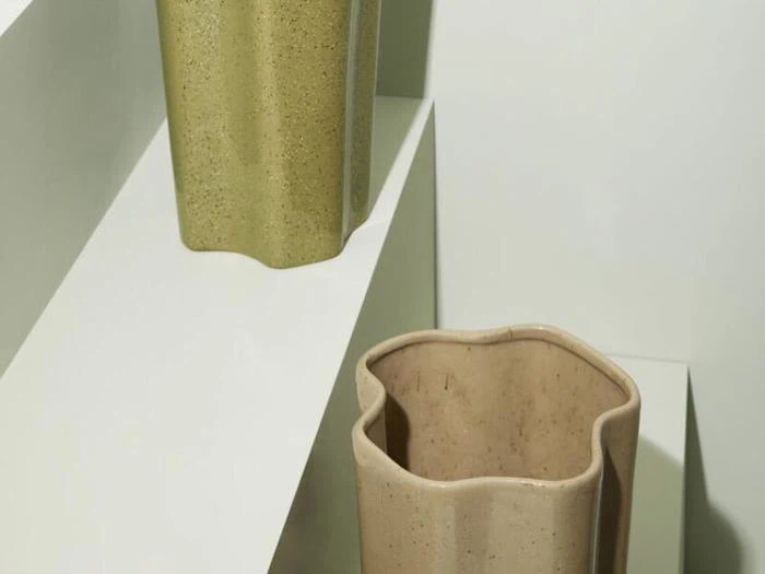 To skulpturelle keramikvaser med bølgede kanter - en olivengrøn og en beige - står på forskudte hvide hylder mod en neutral baggrund.