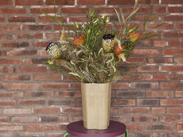 En beige vase med forskellige vilde blomster og grønt står på et lille rundt lilla bord med en murstensvæg som baggrund.