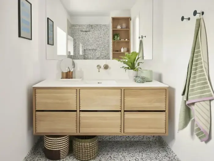 Moderne badeværelse med vaskeskab i lyst træ, hvid bordplade, rundt spejl, grøn plante, håndklæder og kurve nedenunder på et plettet flisegulv. Hylder og vægkroge er synlige.
