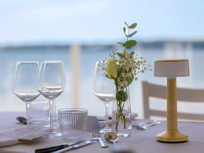 Et bord dækket med glas, bestik, en lille vase med blomster og en moderne lampe med et sløret billede af vand i baggrunden.