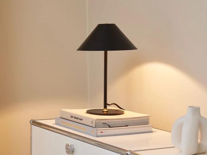 En moderne sort bordlampe sidder på to stablede bøger på et hvidt skab og kaster varmt lys på en beige væg. En hvid pyntegenstand står også på skabet.