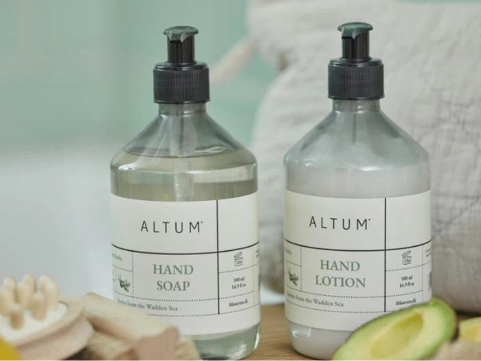 To pumpeflasker med etiketterne ALTUM Hand Soap og ALTUM Hand Lotion står side om side på en flade med en avocado og en blød baggrund i nærheden.