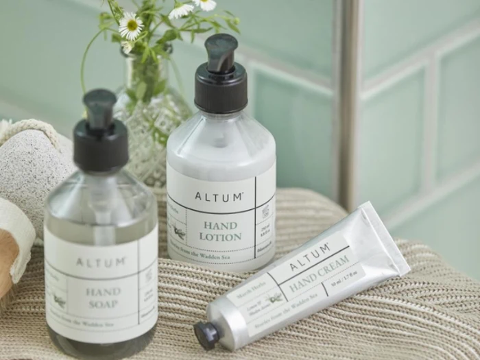 Tre produkter af mærket ALTUM - håndsæbe, håndlotion og håndcreme - står på en badeværelsesdisk med en lille vase med blomster i baggrunden.