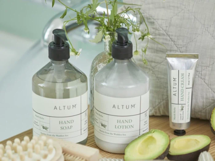 To flasker af mærket ALTUM med mærkerne Håndsæbe og Håndlotion, en tube håndcreme, en avocado og en træbørste på en bakke med blomster i baggrunden.