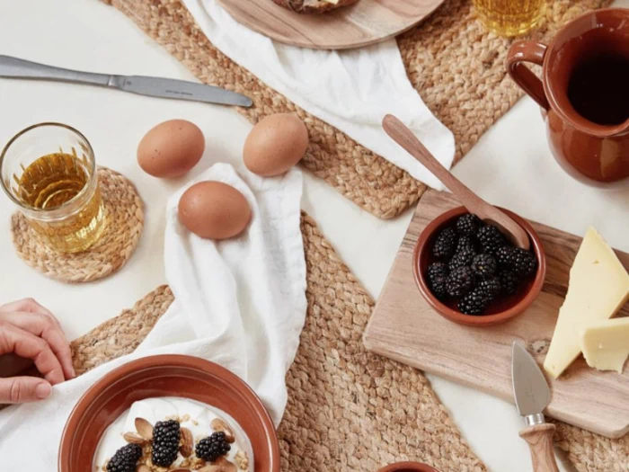 Morgenbordsscene med brombær, ost, æg, et krus, glas med juice og en skål med yoghurt og granola, arrangeret på flettede dækkeservietter.
