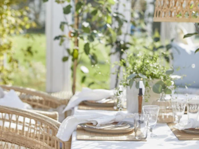 Et solbeskinnet udendørs spisebord dækket med tallerkener, glas og hvide servietter, omgivet af kurvestole og grønne planter.