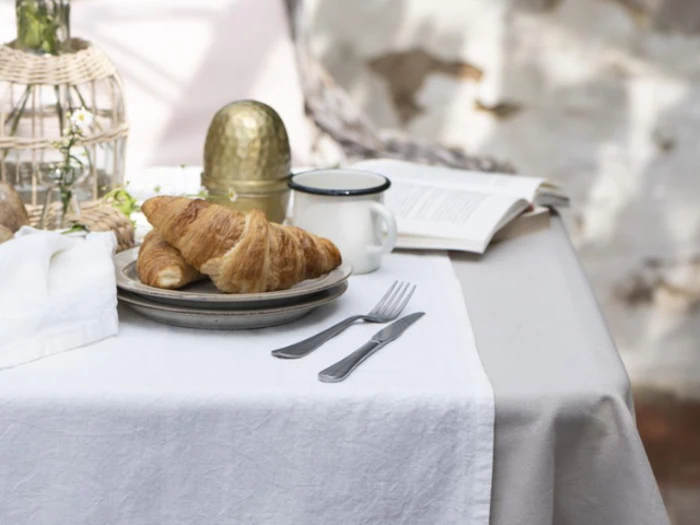 Et bord med to croissanter på en tallerken, et krus, en gaffel og en kniv, et dekorativt æg og en åben bog, alt sammen på en hvid dug.