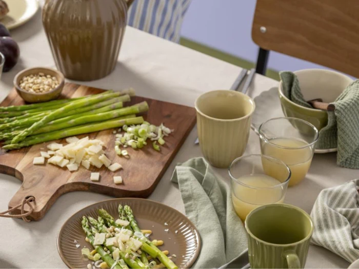 Et bord dækket med asparges, osteterninger og pinjekerner på en træplade, tallerkener med mad, krus, glas med juice og foldede grønne servietter.
